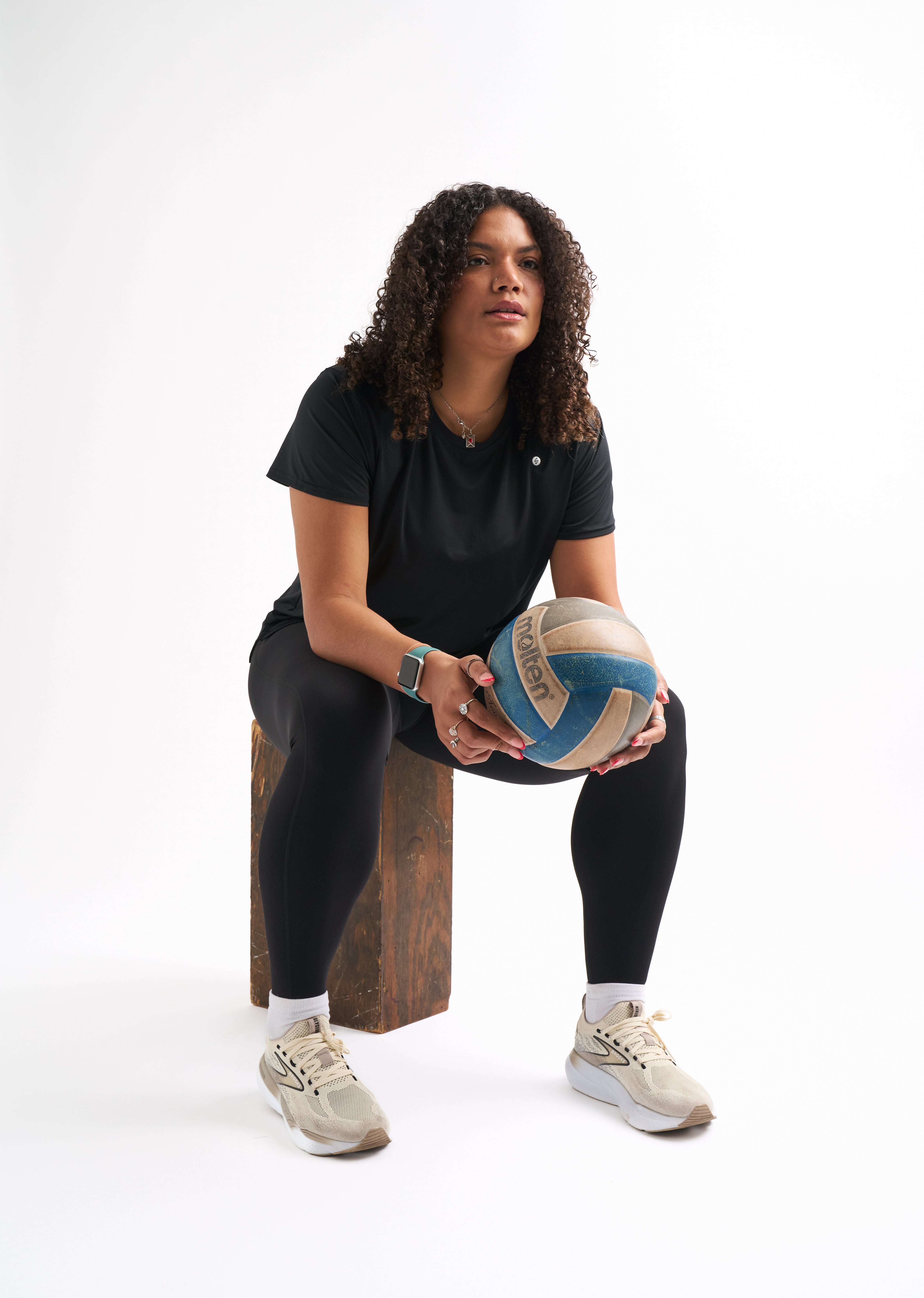 Woman in black activewear sits on a wooden block holding a blue and tan volleyball in both hands.