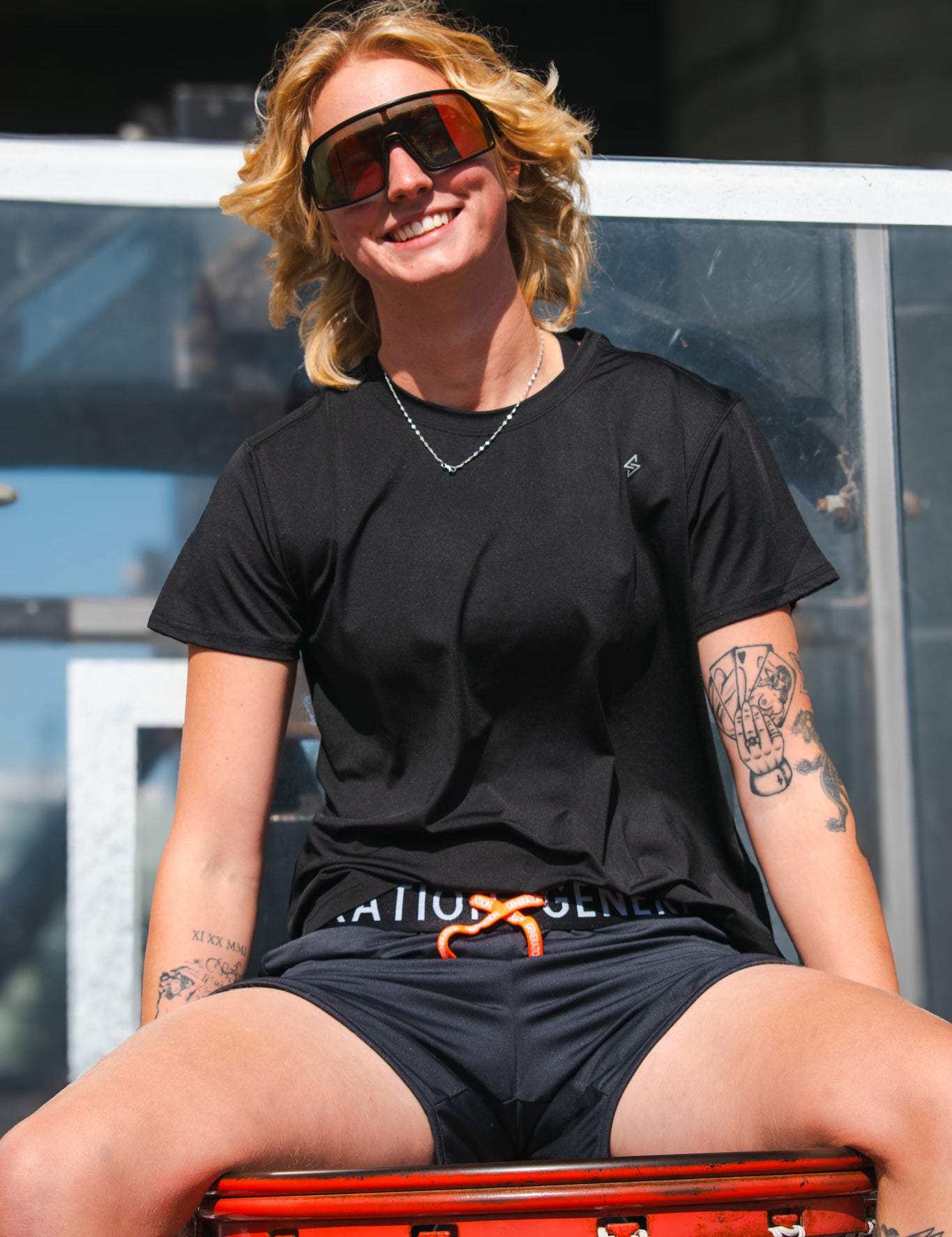 Woman wearing black athletic t-shirt and drawstring running shorts, smiling while sitting on a basketball hoop outdoors