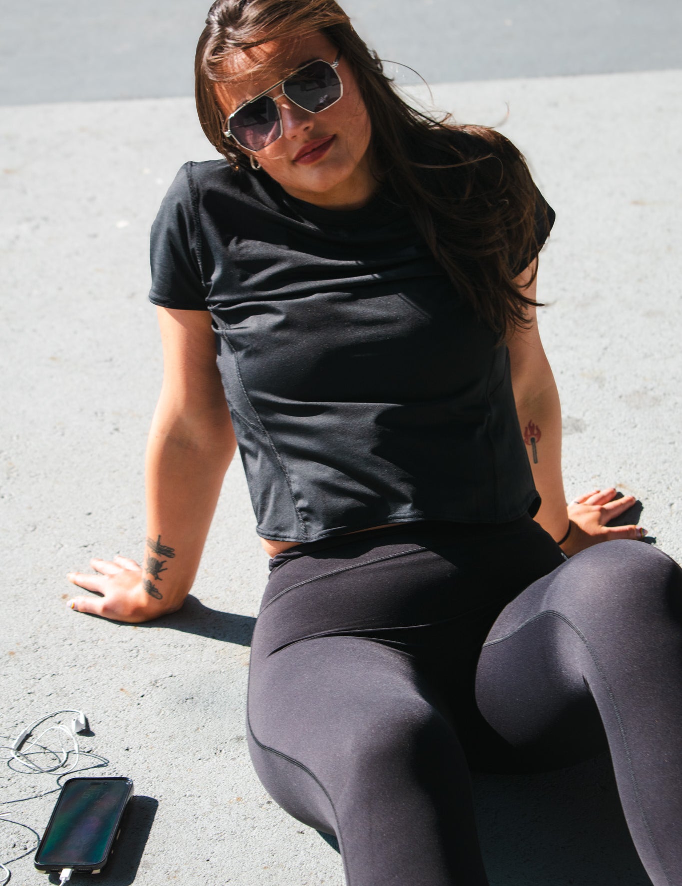 Woman sitting on pavement in sunlight wearing black high-rise leggings and a cropped black workout tee, with phone and earbuds nearby.