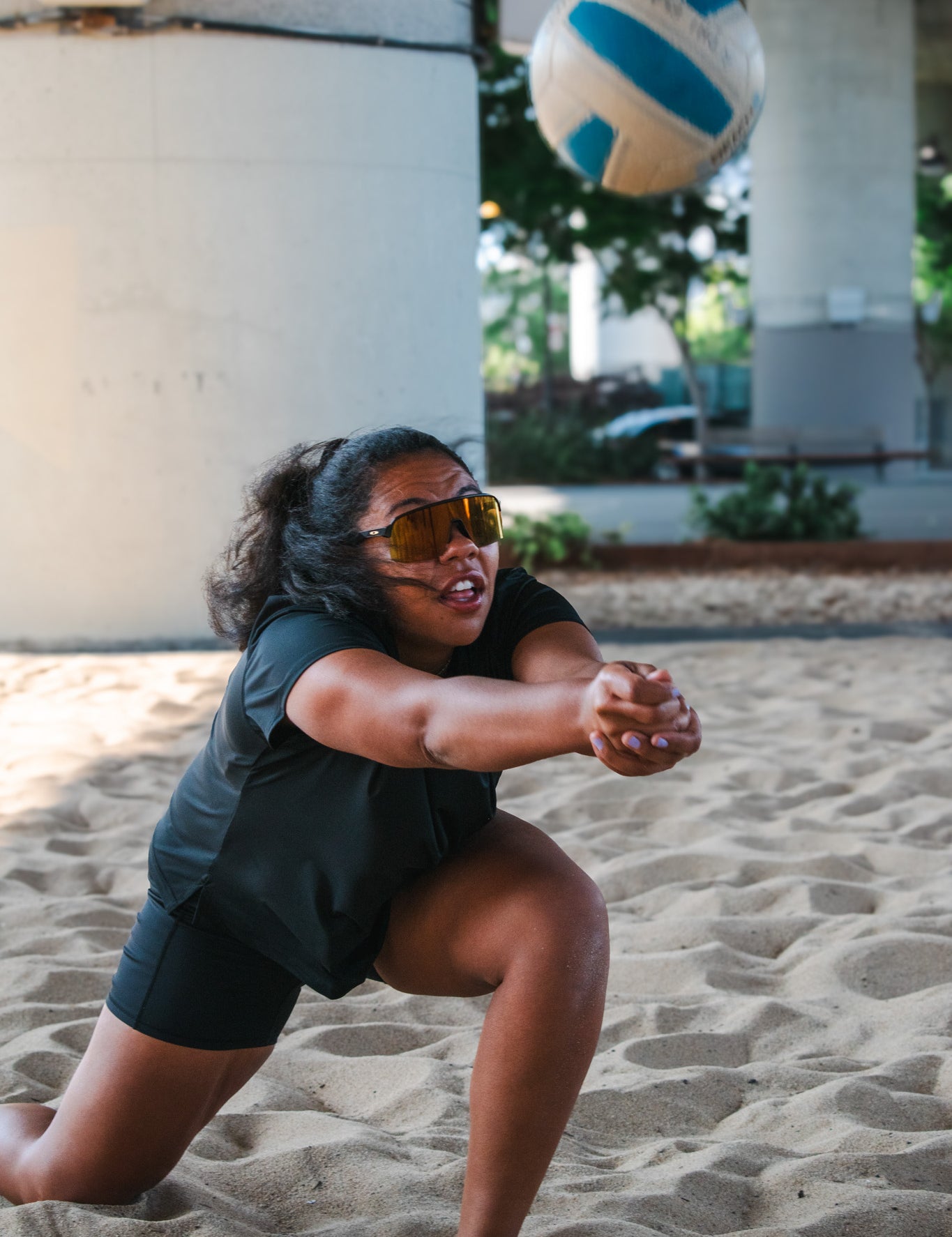 Woman diving in the sand to bump a volleyball while wearing a loose-fit black workout top and biker shorts.