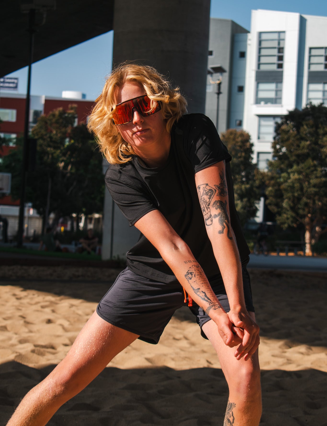Woman in black athletic top and running shorts stretching on a sandy court under an overpass in sunny urban setting.