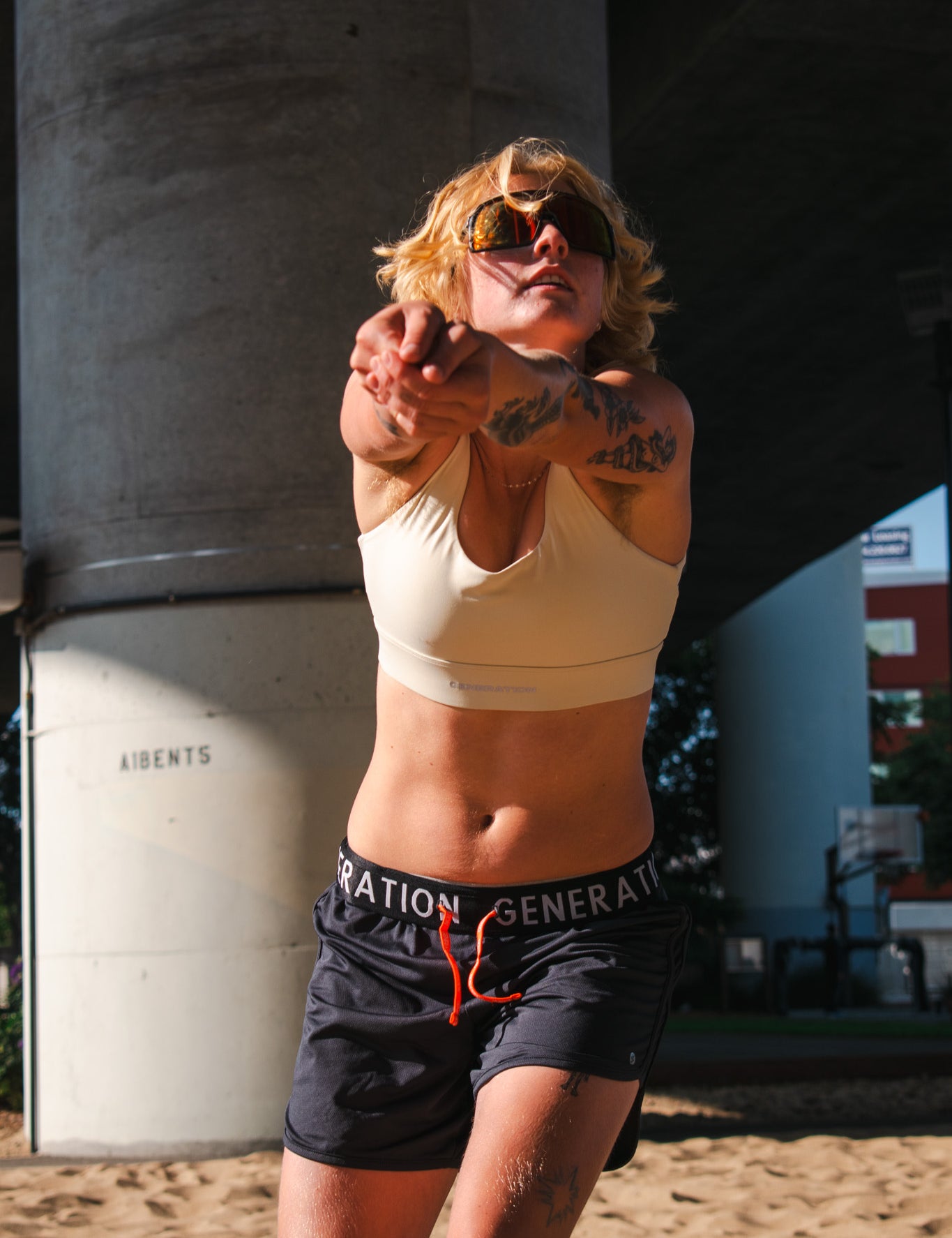 Woman in a beige longline sports bra and drawstring athletic shorts hitting a volleyball on a sand court under an overpass.