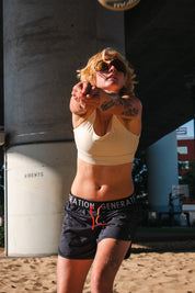 Woman in a beige longline sports bra and drawstring athletic shorts hitting a volleyball on a sand court under an overpass.