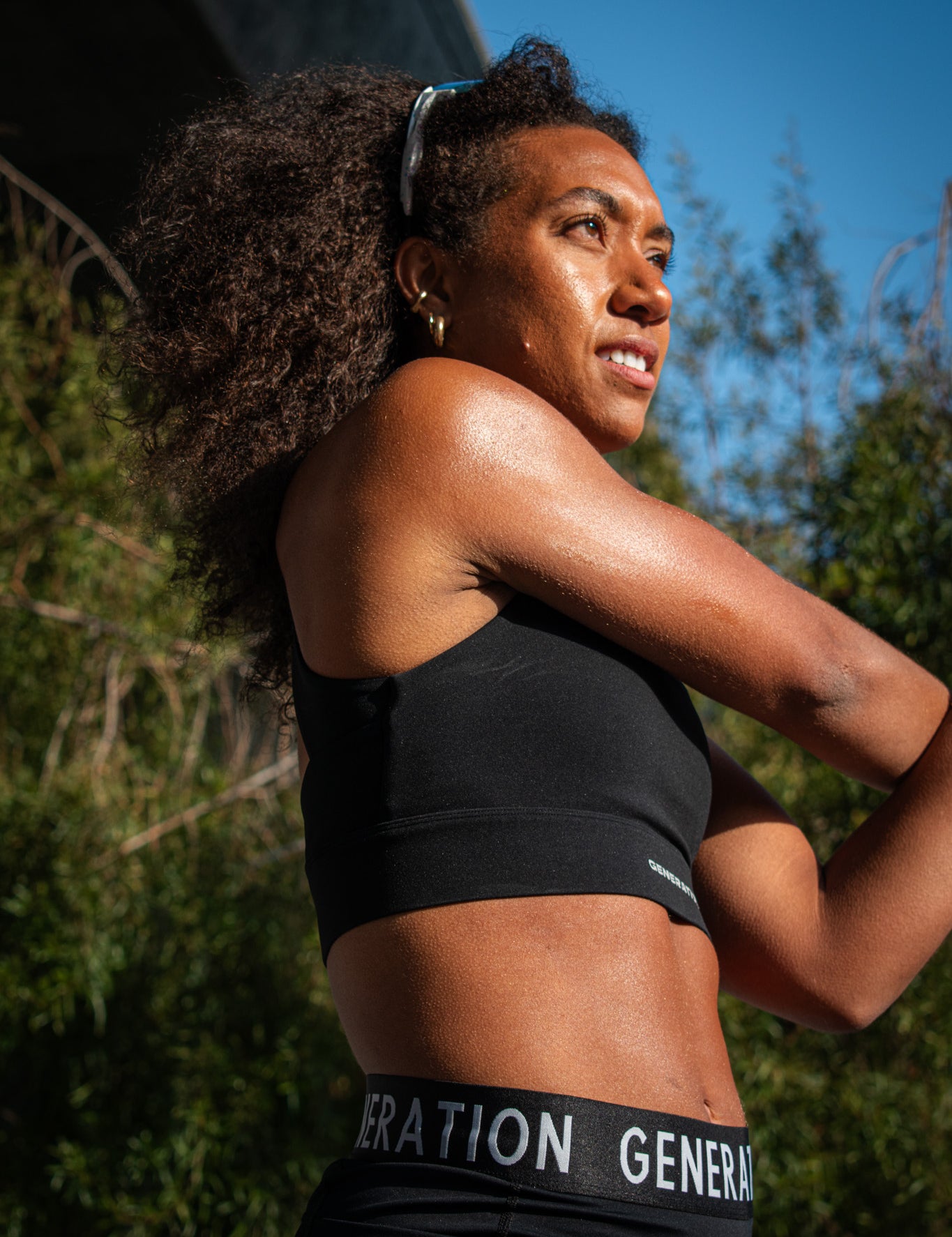 Woman wearing a black sports bra and midrise bike shorts with a logo waistband, stretching outdoors in the sun.