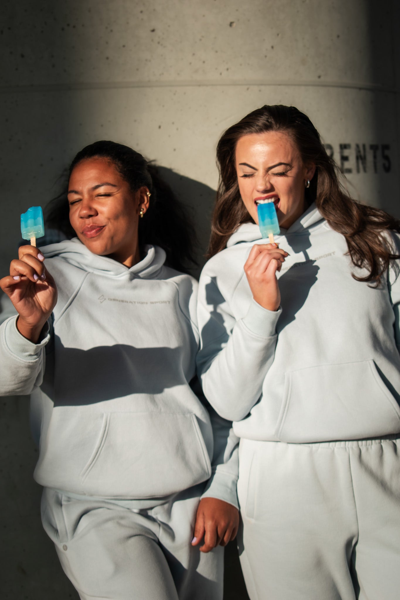 Two people in matching gray sweatsuits holding popsicles against a concrete wall.