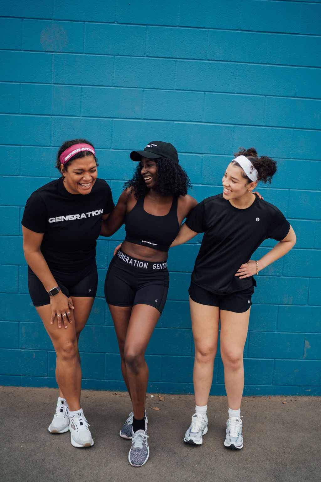 Three women laugh together in performance activewear against a blue wall, showing off sporty energy and team spirit.