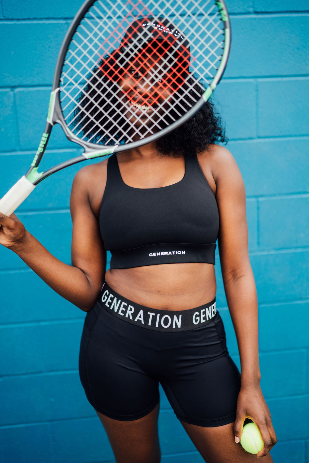 Woman in performance activewear poses with a tennis racket and ball, smiling in front of a bright blue wall.