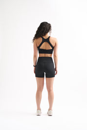 Back view of woman in Assist High Rise Shorts and matching sports bra, standing straight with arms at sides.