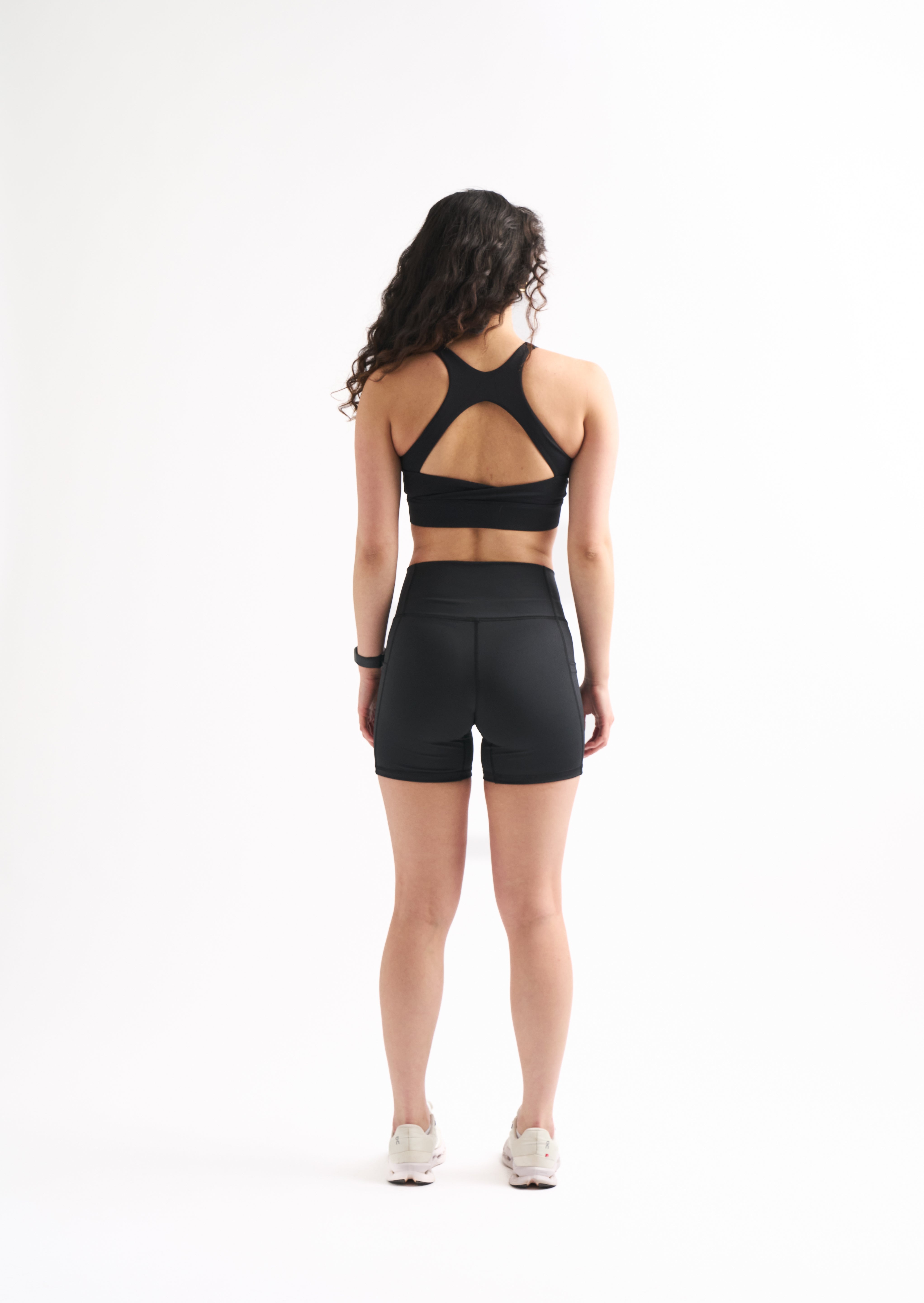 Back view of woman in Assist High Rise Shorts and matching sports bra, standing straight with arms at sides.