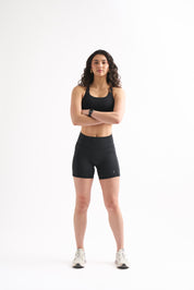 Woman wearing black Assist High Rise Shorts and a sports bra, standing confidently with arms crossed.