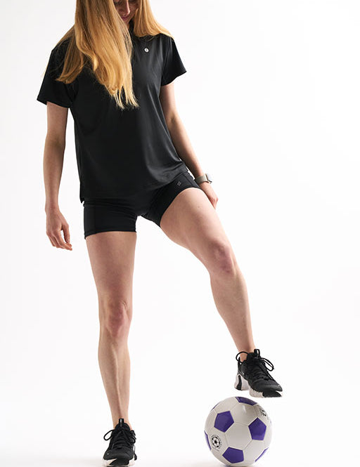 Woman wearing Assist High Rise Shorts and a relaxed black tee with one foot resting on a soccer ball.