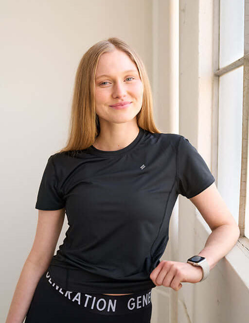 Woman wearing a fitted workout shirt, standing in front of a window smiling.