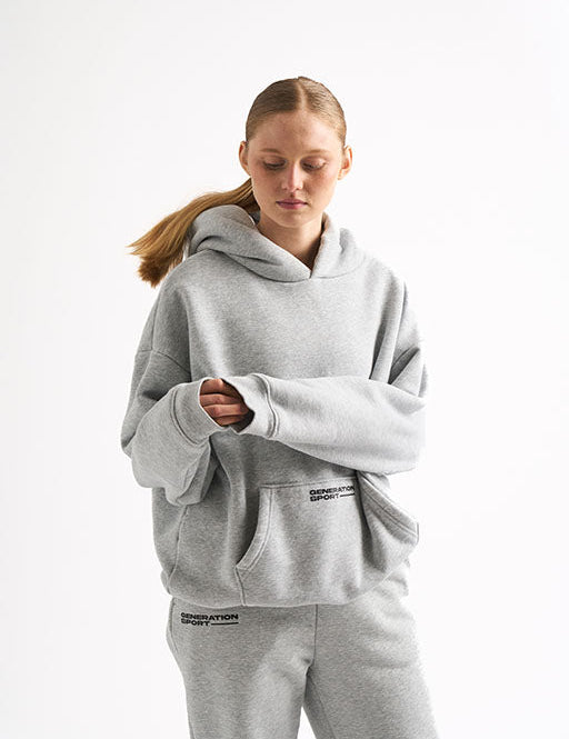 Woman in light gray hoodie and matching joggers, sleeves slightly covering hands, with Generation Sport logo on both pieces.