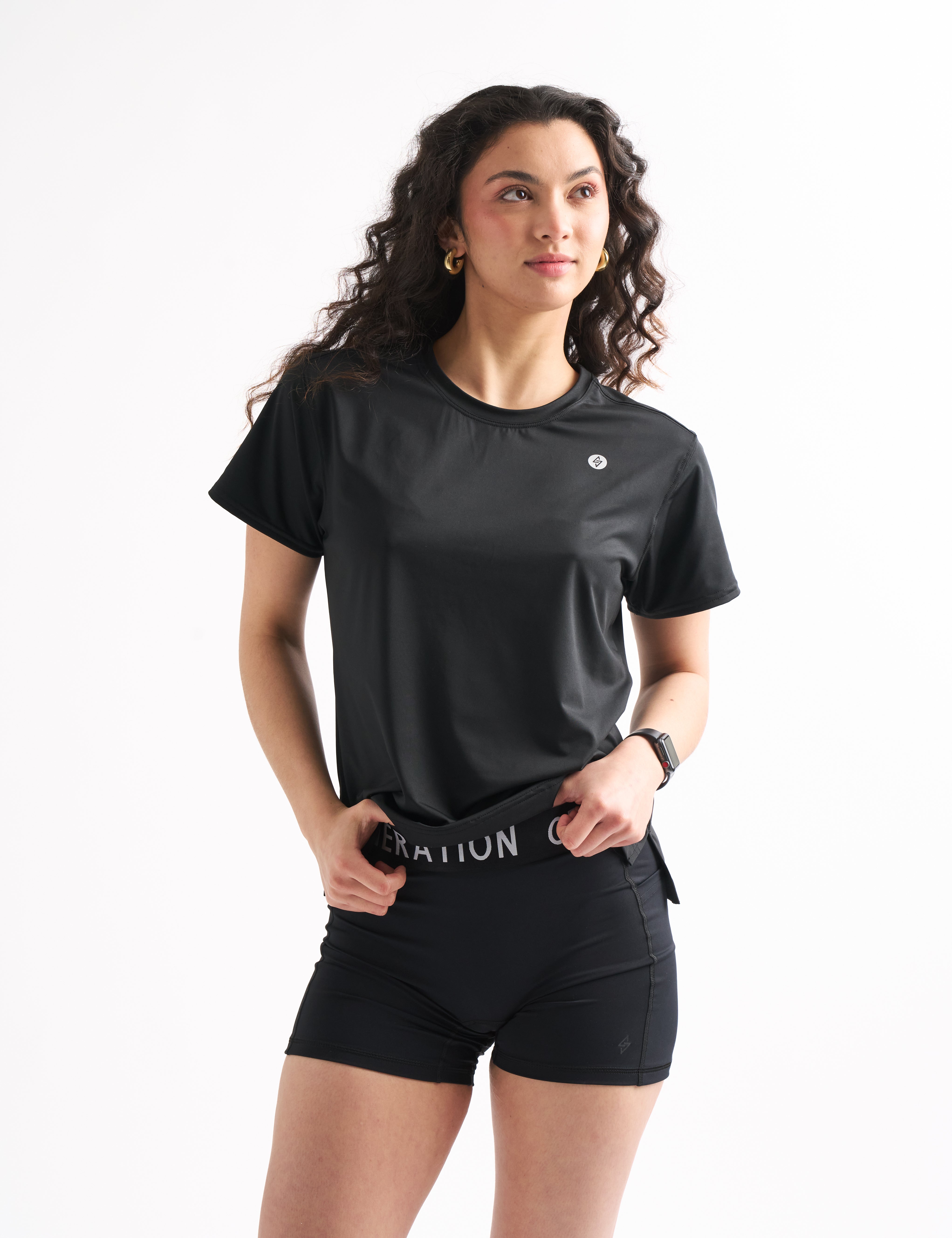 Model slightly lifts black tee to reveal the branded waistband of the Power Moves Midrise Shorts, showcasing a confident, casual look.
