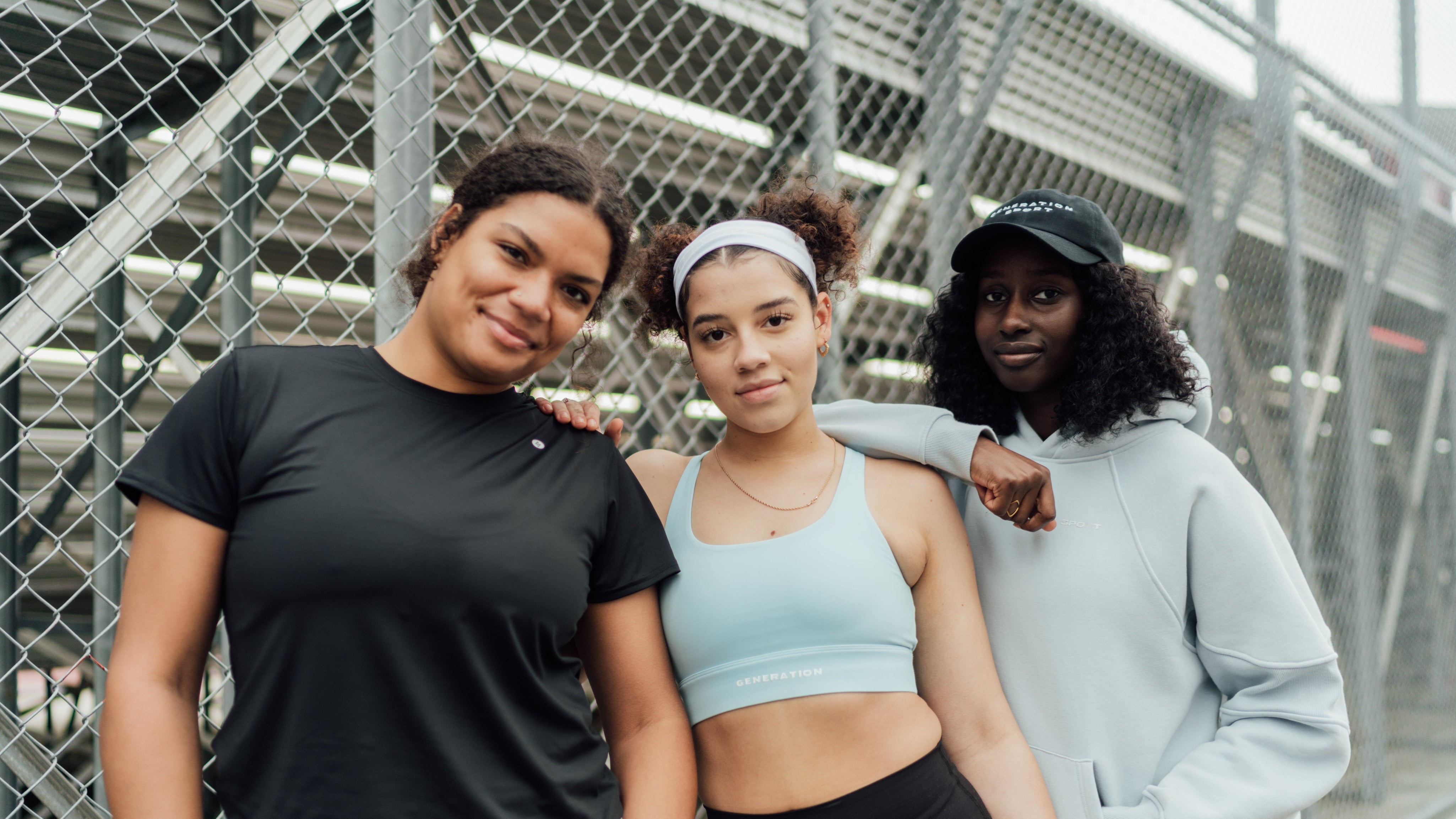 Three women posing casually in women's activewear in front of a chain-link fence, wearing t-shirts, hoodies, and sports bras.