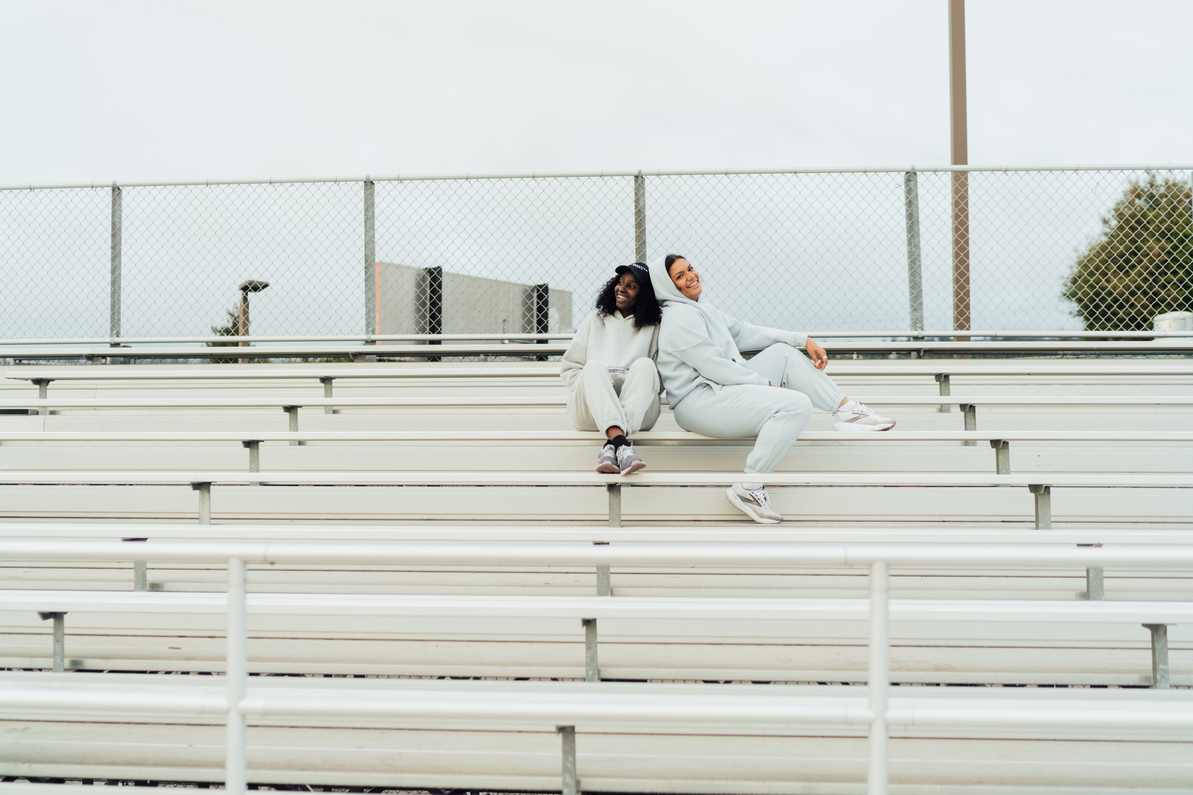 Two women lounging in coordinated grey sweat sets while sitting on bleachers outdoors.