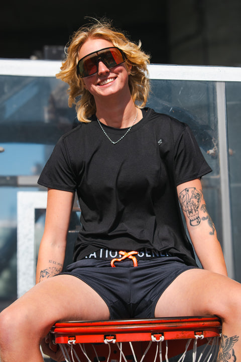 Woman wearing black athletic t-shirt and drawstring running shorts, smiling while sitting on a basketball hoop outdoors