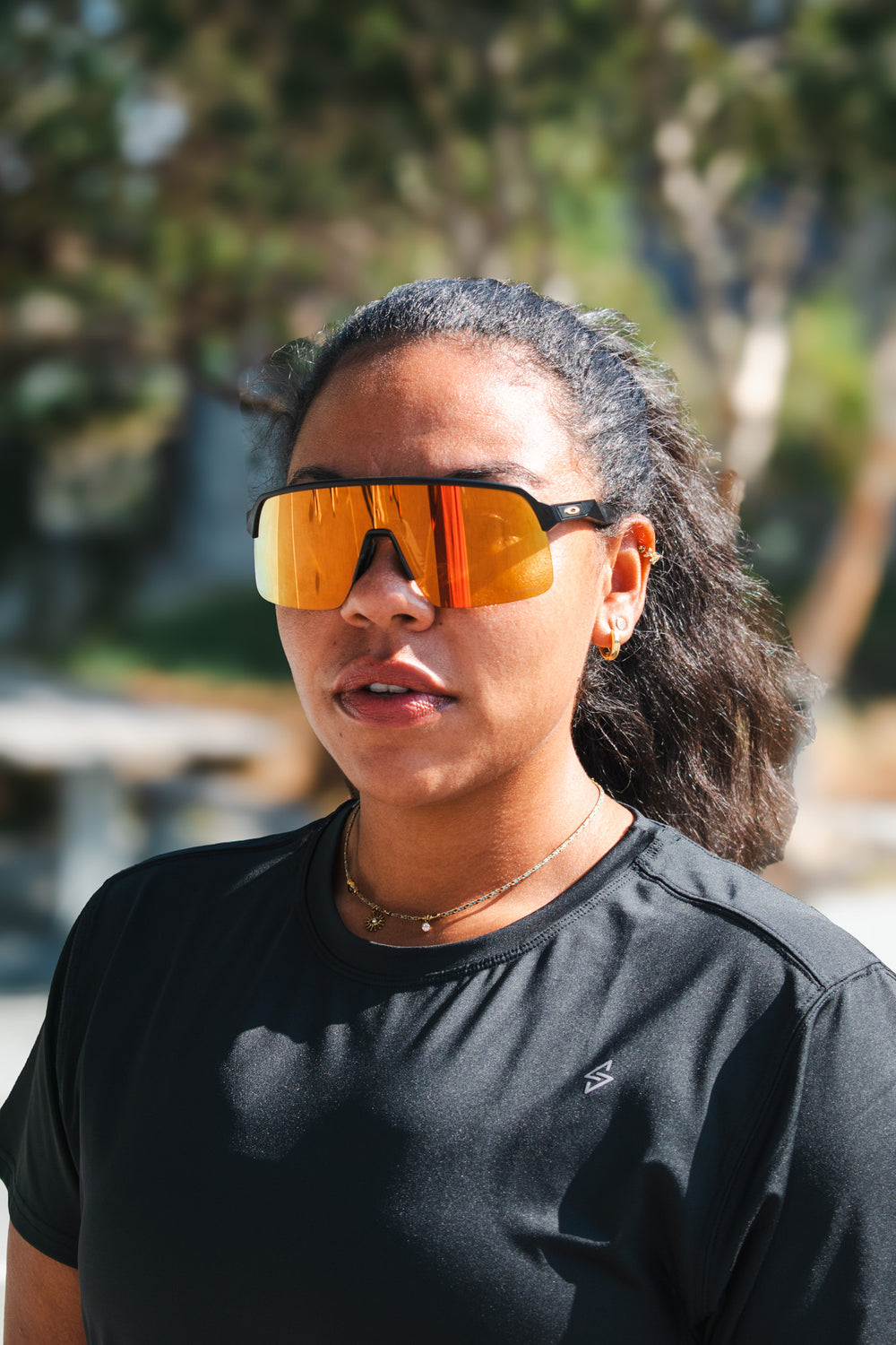 Woman wearing a black relaxed-fit workout top and orange reflective sunglasses, standing outdoors in sunlight.