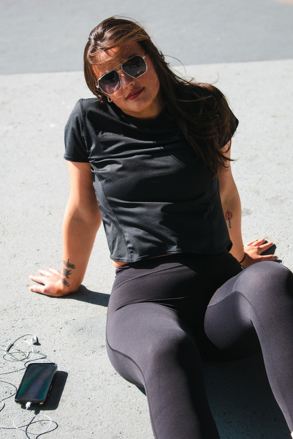 Woman sitting on pavement in sunlight wearing black high-rise leggings and a cropped black workout tee, with phone and earbuds nearby.