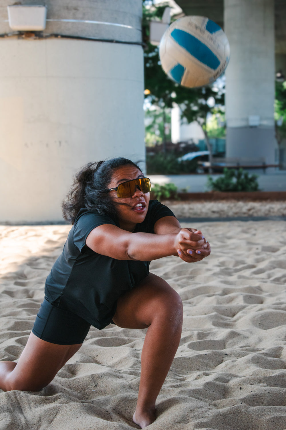Woman diving in the sand to bump a volleyball while wearing a loose-fit black workout top and biker shorts.