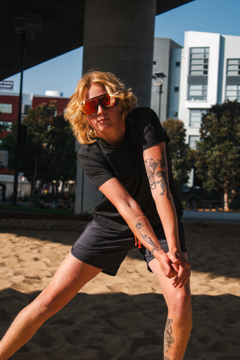 Woman in black athletic top and running shorts stretching on a sandy court under an overpass in sunny urban setting.