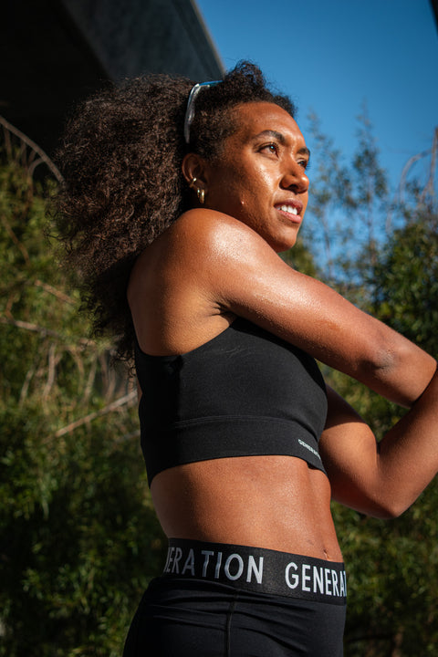 Woman wearing a black sports bra and midrise bike shorts with a logo waistband, stretching outdoors in the sun.