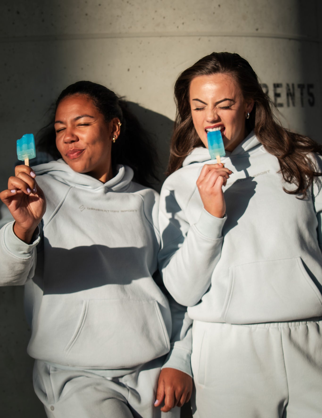 Two people in matching gray sweatsuits holding popsicles against a concrete wall.