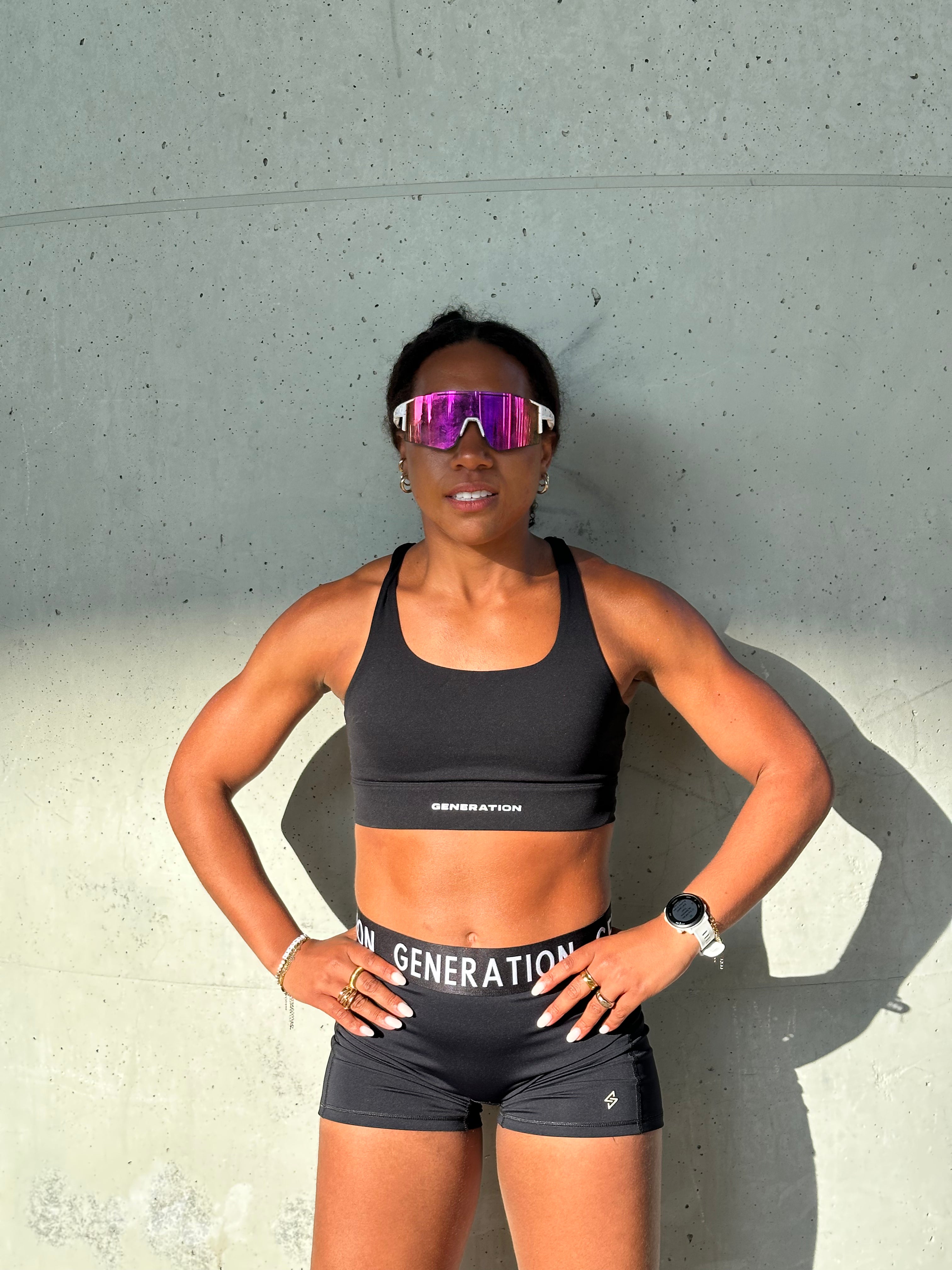 Person wearing a black sports bra and shorts with sunglasses, standing against a concrete wall.