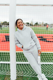 Woman in a light gray fleece hoodie and joggers smiling and leaning against a soccer goalpost on a track field.