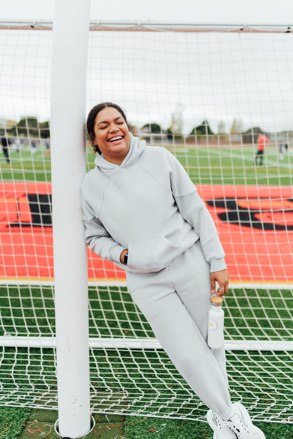 Woman in a light gray fleece hoodie and joggers smiling and leaning against a soccer goalpost on a track field.