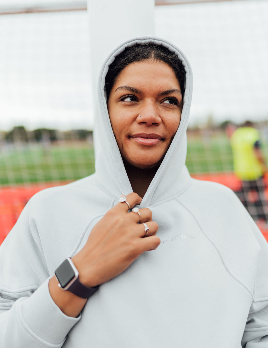 Woman wearing a light gray fleece hoodie with the hood up, standing on a field and looking to the side with a slight smile.