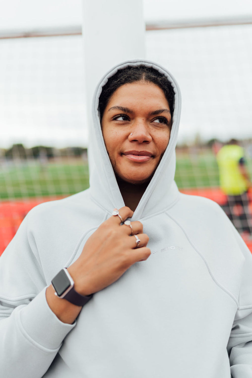 Woman wearing a light gray fleece hoodie with the hood up, standing on a field and looking to the side with a slight smile.