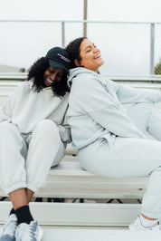 Two women in matching fleece jogger sets sitting on bleachers, smiling and leaning on each other in relaxed postures.
