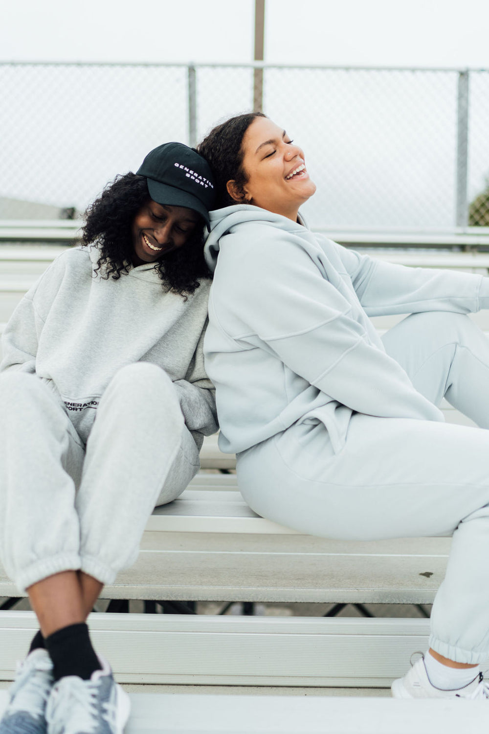 Two women in matching fleece jogger sets sitting on bleachers, smiling and leaning on each other in relaxed postures.