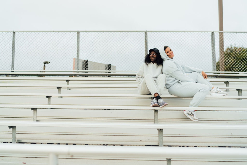 Two women in cozy sweat sets lounging on bleachers in light gray hoodies and sweatpants, smiling and relaxed.