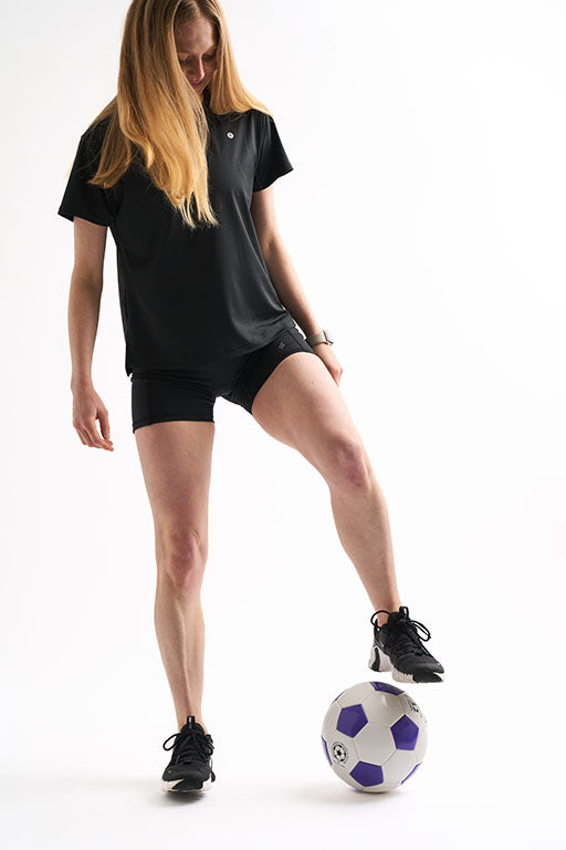 Woman wearing Assist High Rise Shorts and a relaxed black tee with one foot resting on a soccer ball.