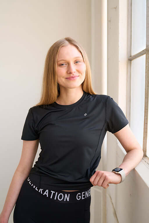Woman wearing a fitted workout shirt, standing in front of a window smiling.