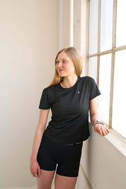Woman in fitted black workout shirt, standing in front of a window and looking away.