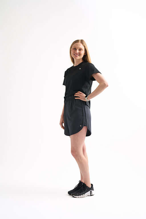 Side view of woman wearing Locked In running shorts and black athletic shoes, standing with hand on hip and smiling.