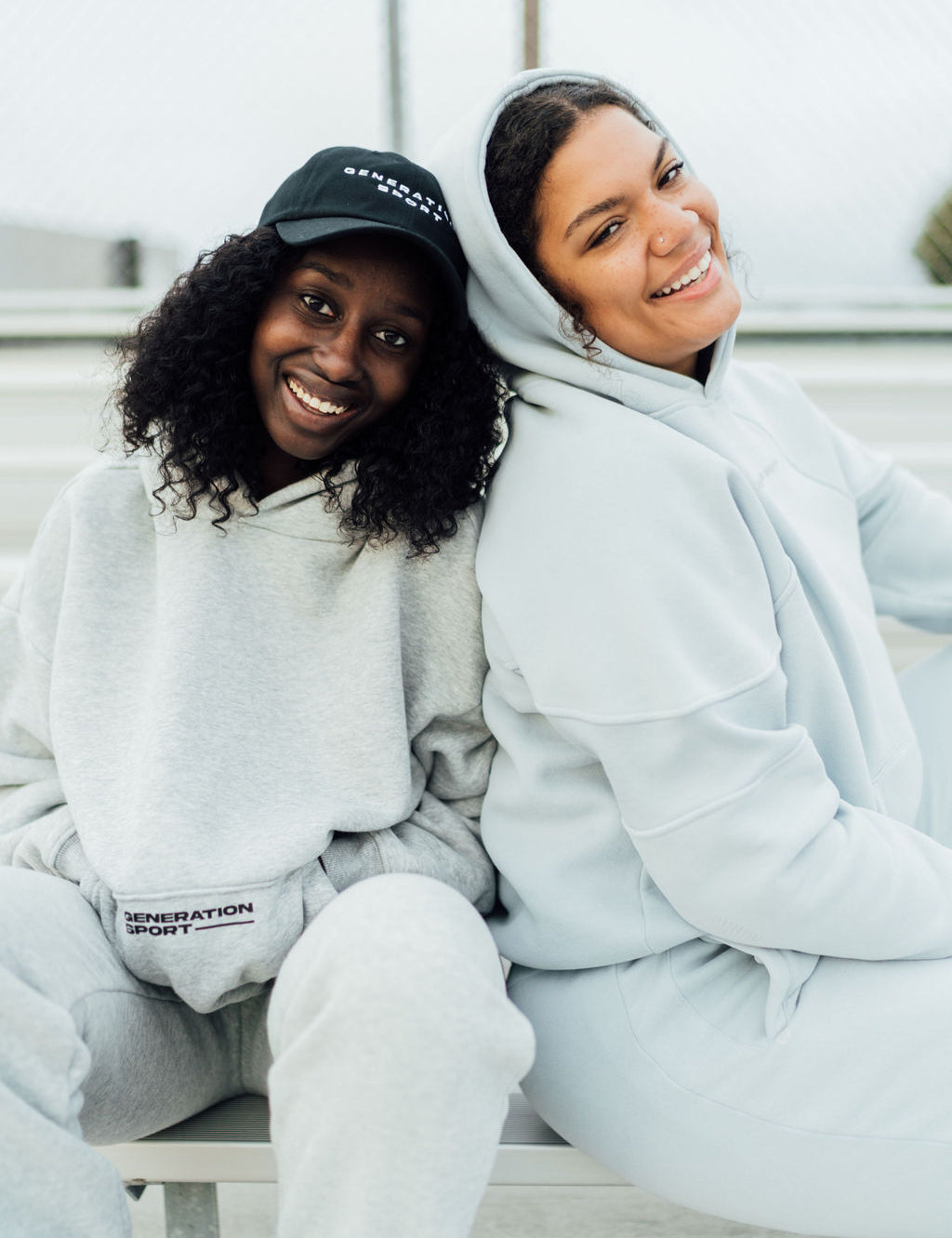 Two women sitting on bleachers wearing Generation Sport classic fleece-lined hoodies in gray and soft blue