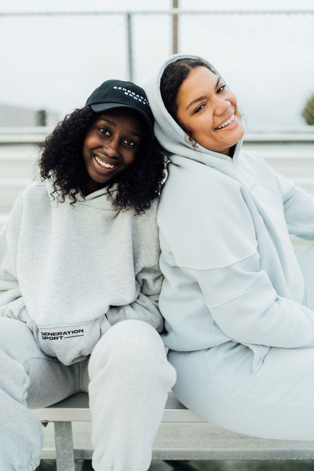 Two women sitting on bleachers wearing Generation Sport classic fleece-lined hoodies in gray and soft blue