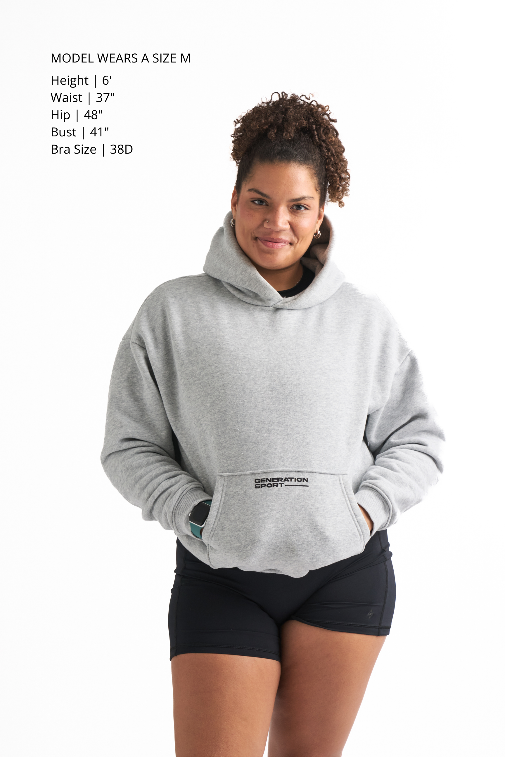 Woman wearing a light gray classic fleece-lined hoodie with black workout shorts, hands in the hoodie pocket and smiling
