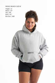 Woman wearing a light gray classic fleece-lined hoodie with black workout shorts, hands in the hoodie pocket and smiling