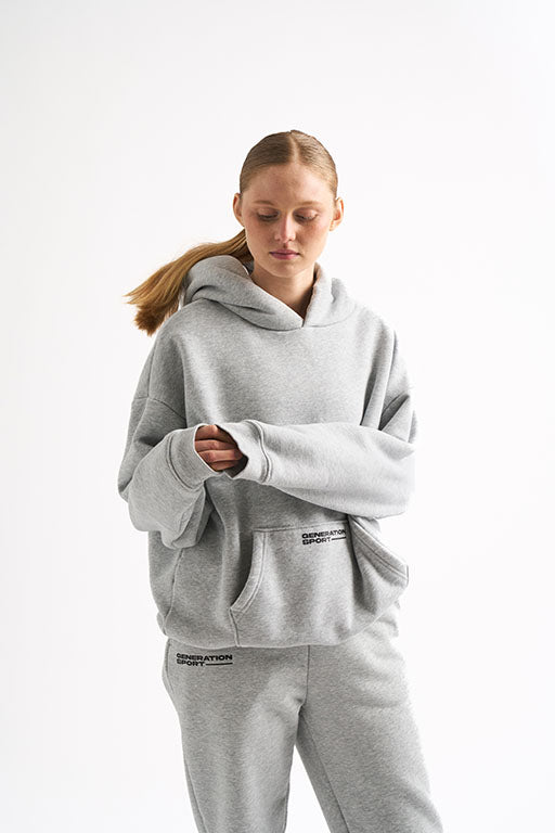 Woman in light gray hoodie and matching joggers, sleeves slightly covering hands, with Generation Sport logo on both pieces.