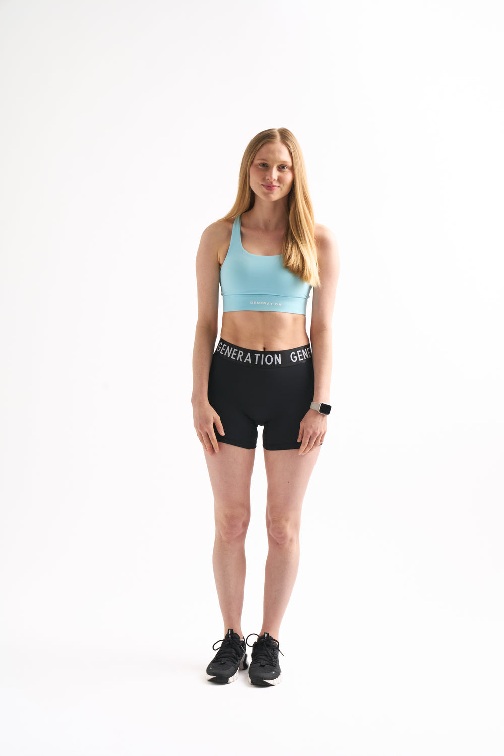 Model stands in a light blue sports bra and Power Moves Midrise Shorts, showing the relaxed fit and branded waistband. 