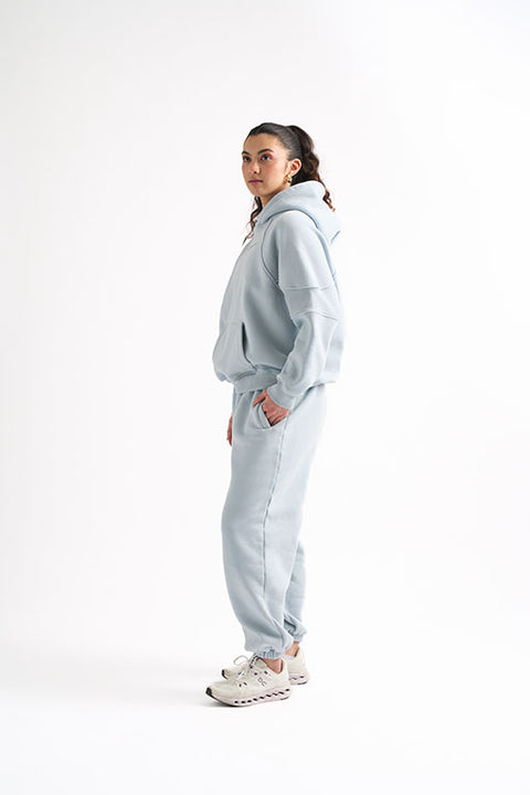 Side view of woman wearing a light blue oversized sweat set, standing with one hand in her pocket and looking ahead.