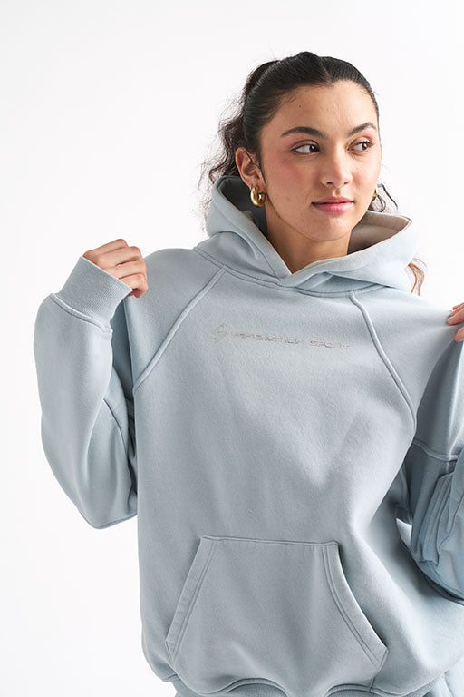 Woman holding the collar of her light blue oversized hoodie, showing the embroidered Generation Sport logo on the chest.
